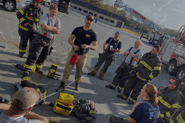 Firefighters gather outdoors for hands-on training beside a fire engine and ladder truck.