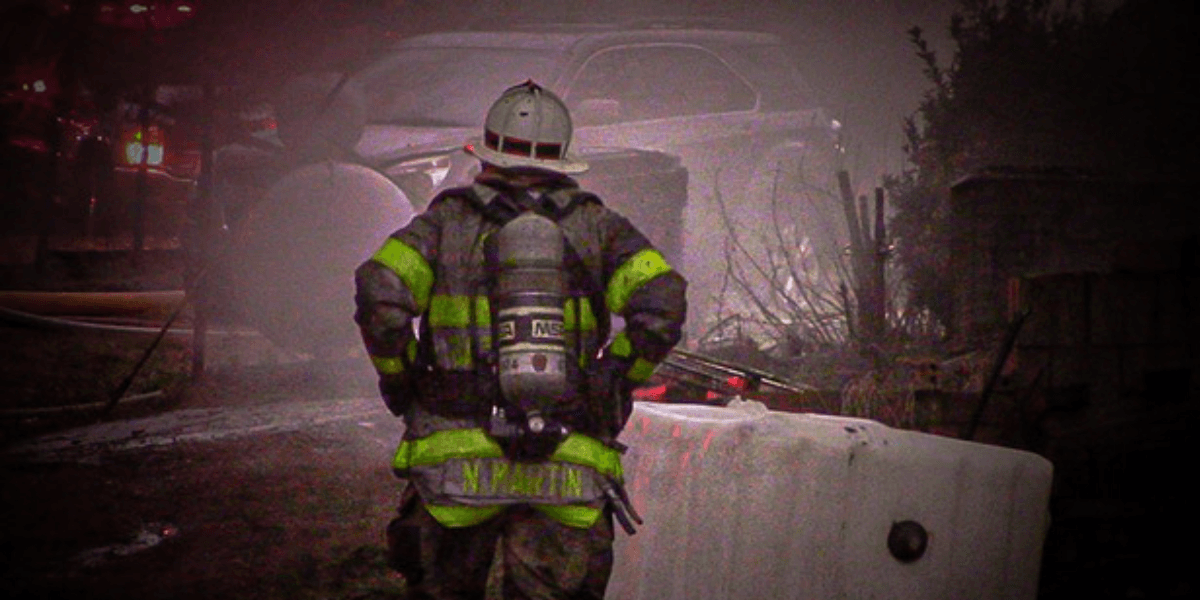 Firefighter in protective gear stands near a fire engine at a smoky nighttime fire scene.