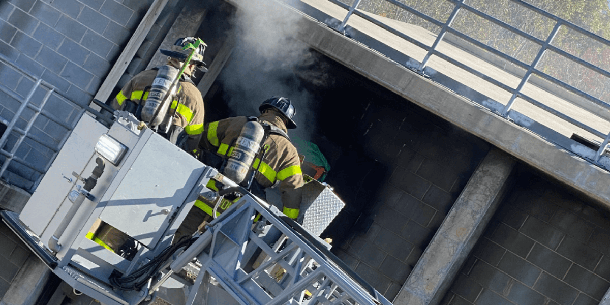 Two firefighters on an aerial platform approach a smoke-filled building opening.